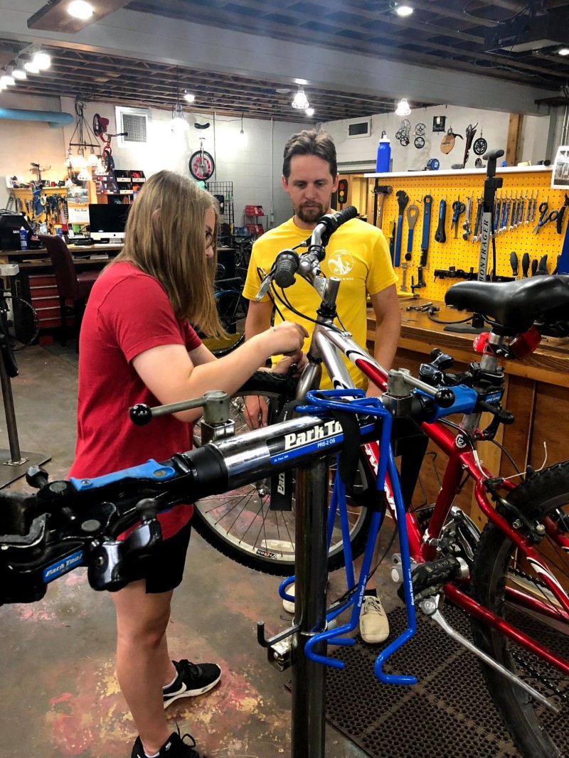 Two people walking and working on a bicycle in a work stand with tools in a nearby workstation