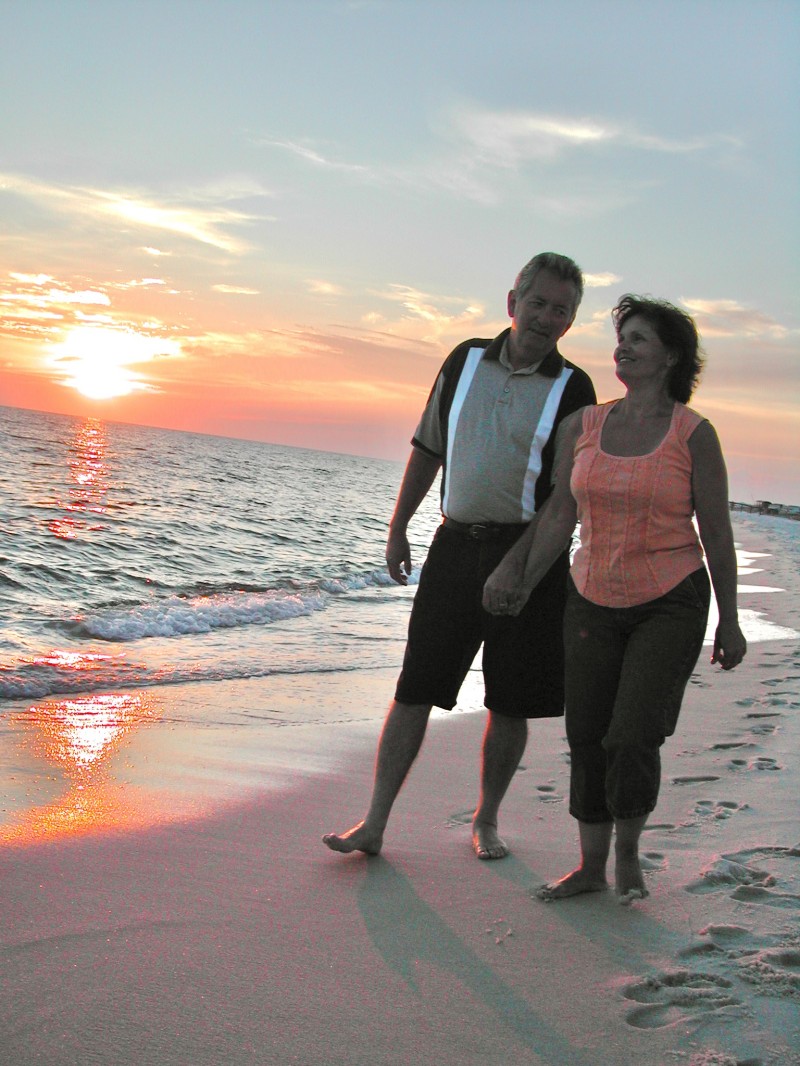 2 people, a man and woman holding hands and walking along the beach at sunset